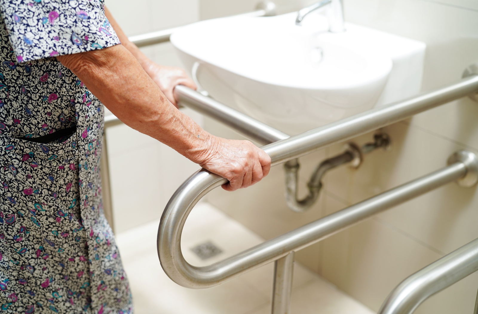 A senior in a colourful print holds onto a grab bar while using the sink in a bathroom