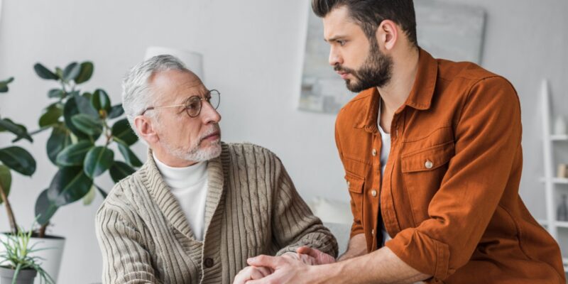 Adult son holding hands with elderly father in a moment of support at home, reflecting emotional connection and caregiving.