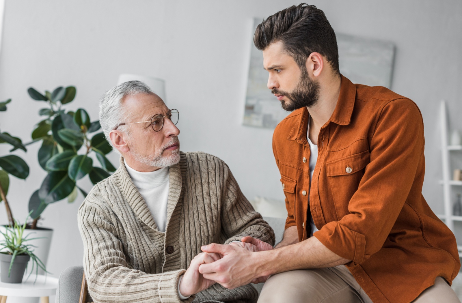 Adult son holding hands with elderly father in a moment of support at home, reflecting emotional connection and caregiving.
