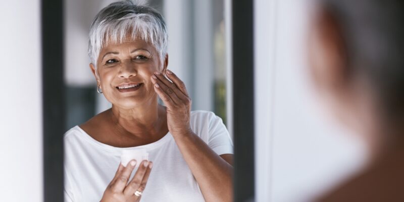 An older adult smiles while carefully applying moisturizer to their cheek in front of a mirror