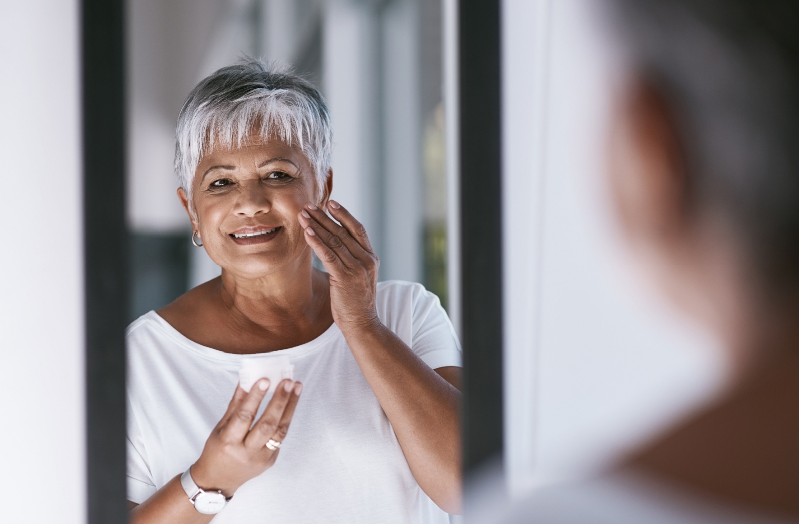 An older adult smiles while carefully applying moisturizer to their cheek in front of a mirror
