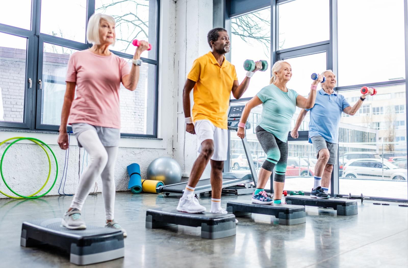 A group of senior friends enjoy a light mobility workout class together in a brightly-lit fitness studio.