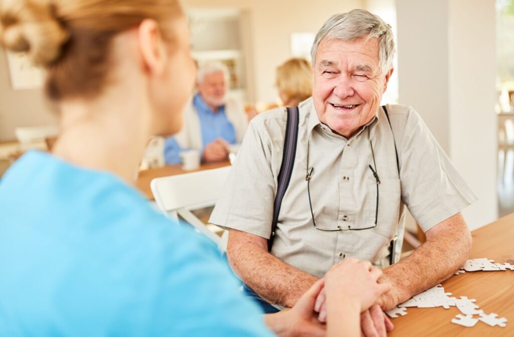 A senior smiles while a caregiver grasps their hands as they work on building a puzzle.