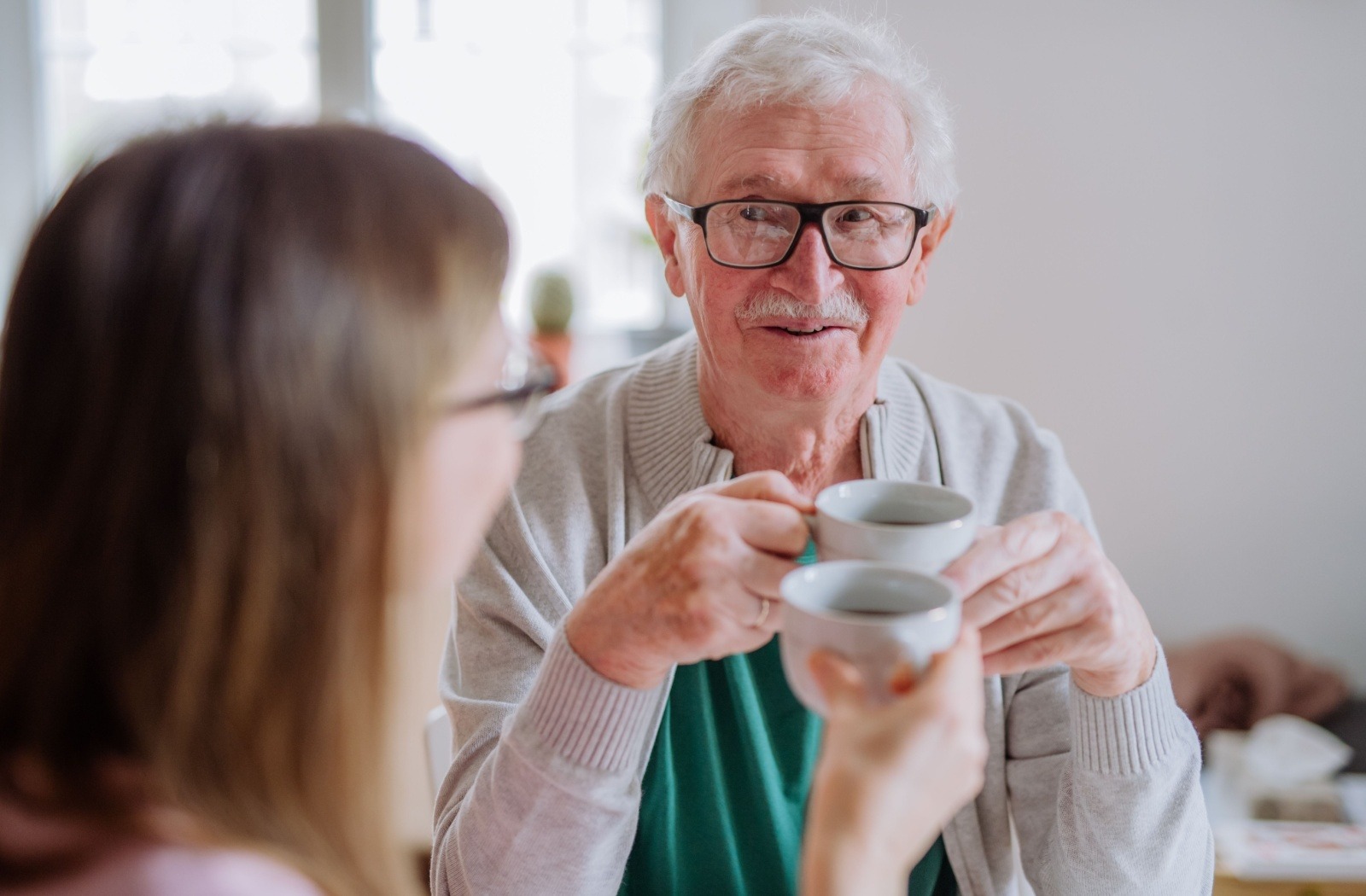 Older man with glasses smiling, holding two coffee cups in a bright room while sharing a coffee with a friend.