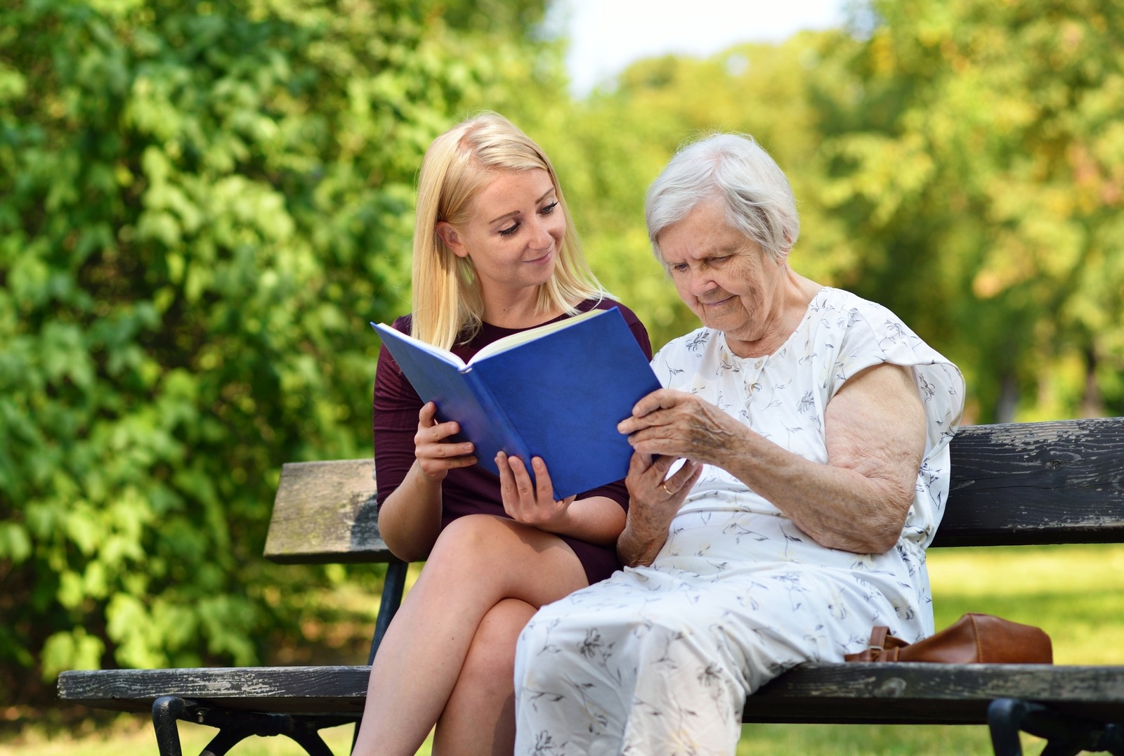 Young caregiver reading a book to an elderly woman in the park, sitting on a bench on a bright afternoon