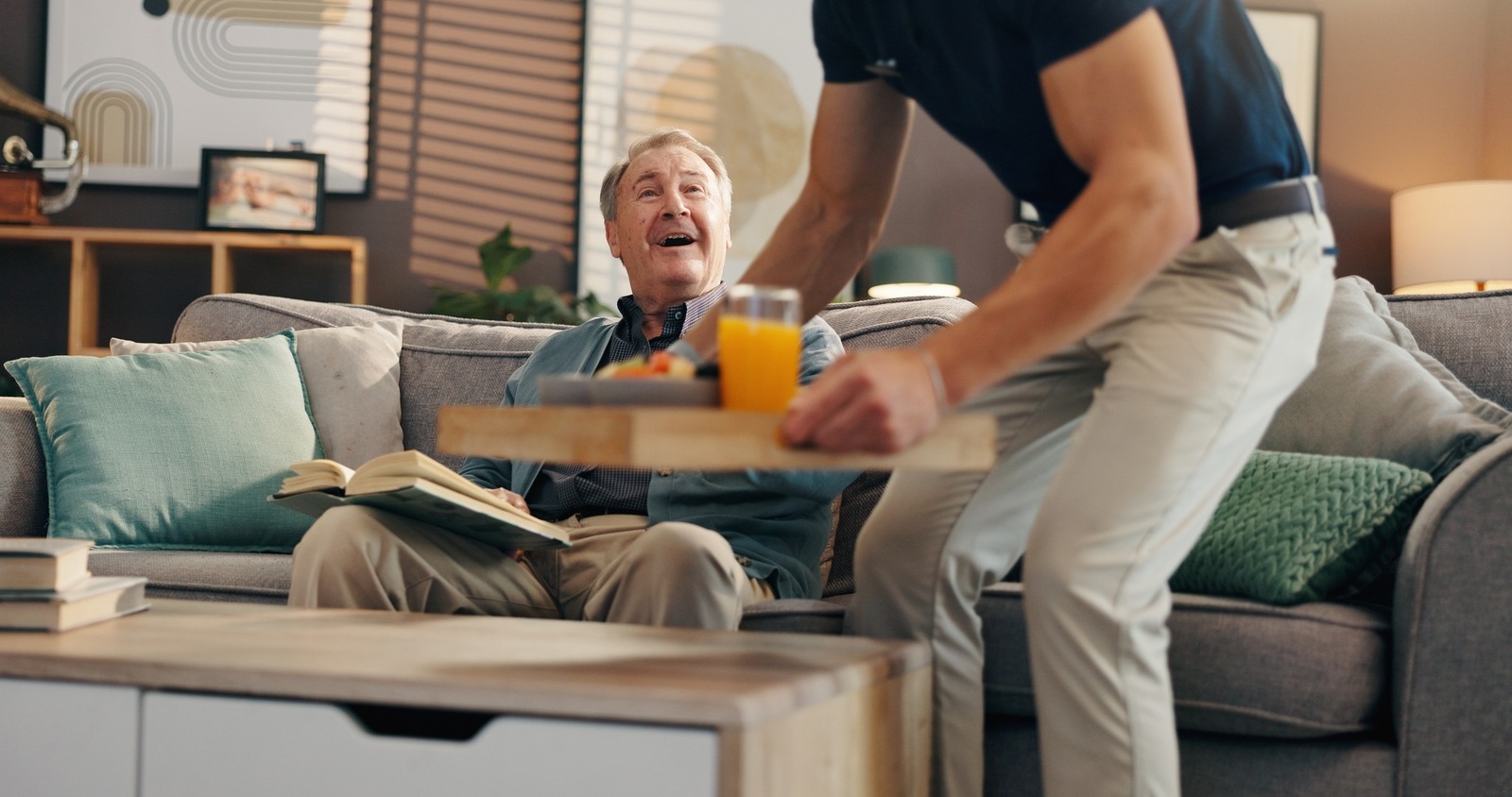Caregiver with food tray for an elderly man reading a book happily