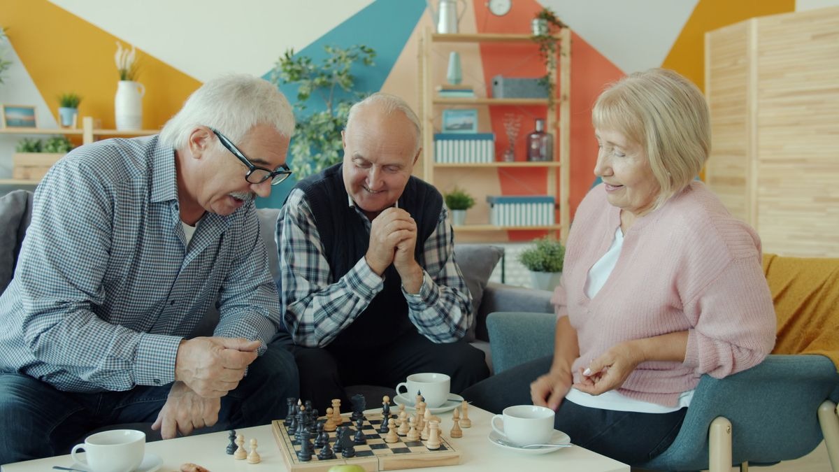 Three older adults smiling around a coffee table with a chessboard and cups of tea in a brightly decorated living room.